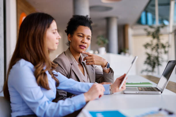 Two businesswomen are sitting at a desk in a modern office, collaborating on a project using a digital tablet and laptop, discussing strategies and analyzing data