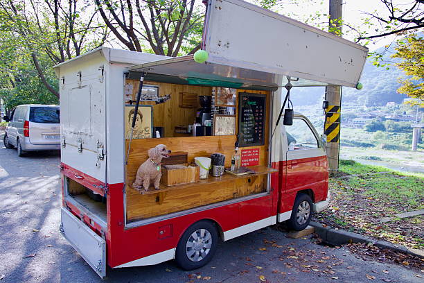 Gurye County, South Korea - October 3rd, 2021: A small mobile coffee truck parked along a scenic riverside road serves fresh drinks to passing cyclists and travelers, with a friendly poodle sitting on the counter.