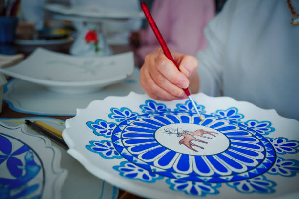 A close-up view of an artist painting a decorative ceramic plate with intricate yellow floral patterns in a pottery studio. The workspace is filled with various brushes, colorful paints, and ceramic pieces, showcasing the artistic process and creativity involved in hand-painting ceramics. In the background, other artisans work on their ceramic projects, contributing to the collaborative and inspiring atmosphere of the studio. This image highlights the craftsmanship and dedication required for traditional ceramic art.