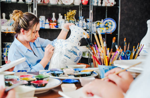 In a ceramics workshop, a woman is drawing hand-painted designs on a large vase. she is painting  carefully. The workshop environment includes ceramic products and painting materials. A scene reflecting the themes of art, craft, handicraft, ceramic painting, women's solidarity and creativity.