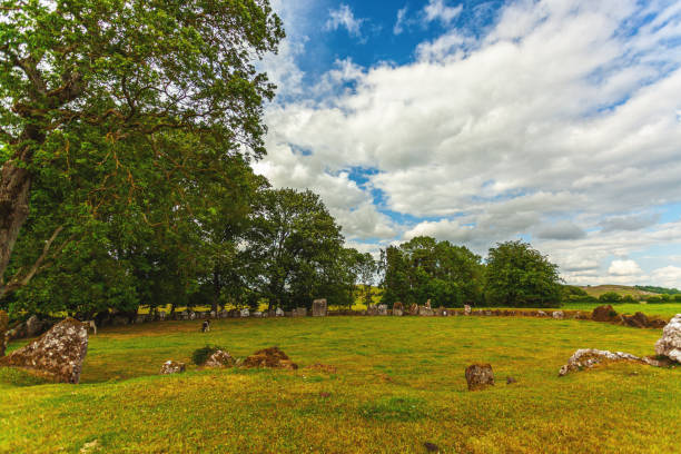 the Grange Stone Circle, an ancient megalithic site in Ireland. The scene features a large, green field with standing stones arranged in a circle. Trees surround the field, adding to the natural setting. The sky is bright with white, fluffy clouds, creating a dynamic backdrop. The image conveys a sense of history and mystery, highlighting the ancient heritage of the irish landscape. County Limerick, Ireland