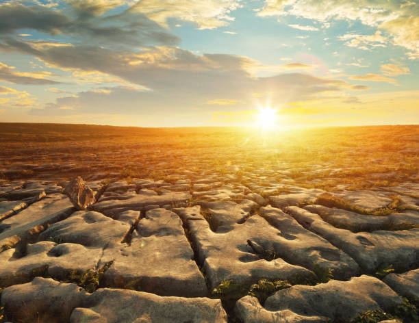 sunset view of the Burren in County Clare, Ireland, featuring a unique karst landscape. The foreground showcases the characteristic limestone pavement with its distinctive cracks and weathered rocks. A solitary rock stands out against the textured terrain. The setting sun is visible on the horizon, casting a warm, golden light over the landscape. The sky is filled with scattered clouds, adding to the dramatic atmosphere.