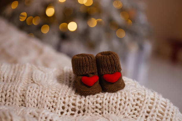 A pair of brown knitted baby booties with red heart decorations placed on a soft white knitted blanket. A blurred Christmas tree with warm lights in the background. Concept warmth, love, and family.