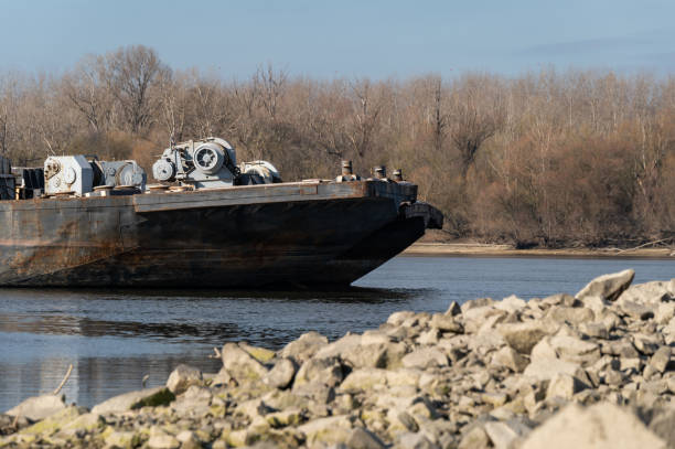 A close-up view of a rusty cargo barge with industrial machinery on its deck, floating on a river near a rocky shoreline. The worn metal surface and mechanical equipment highlight the vessel's industrial purpose against a backdrop of leafless trees.