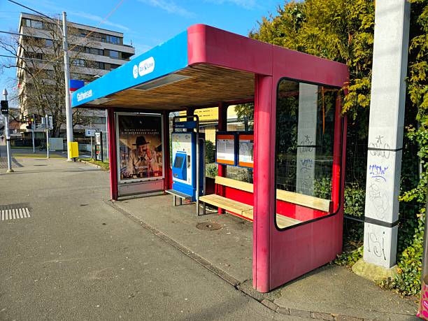 Wide shot of two senior female friends waiting inside a bus shelter in Newcastle-upon-Tyne, North East England. They are both wearing warm winter coats and eyeglasses, looking off-frame as they wait for a bus, one holding a net bag filled with fruit. One of the females is living with dementia.\n\nVideos similar to this scenario available.