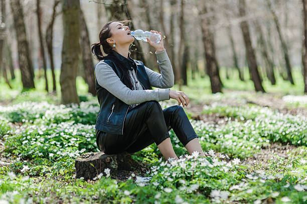 istock A black-haired woman drinking water from a plastic bottle in a spring forest and sitting on a tree stump 2205398735