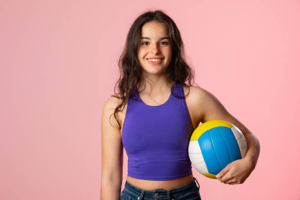 Young woman with long brown hair smiles while holding a volleyball, showcasing her passion for the sport against a vibrant pink backdrop, ready for a game or practice