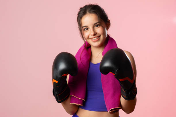 Portrait of a smiling young woman, wearing boxing gloves and a towel draped around her neck, expressing her passion for boxing and fitness while radiating confidence and determination