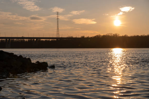A scenic view of a river at sunset, with the sun casting a golden reflection on the rippling water. A bridge and a towering power line structure silhouette against the warm hues of the sky.