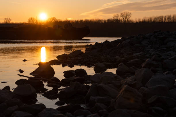 A golden sunset casts a warm glow over a rocky riverbank, reflecting off the calm water. In the background, a silhouetted cargo ship moves along the river, creating a serene yet industrial contrast.