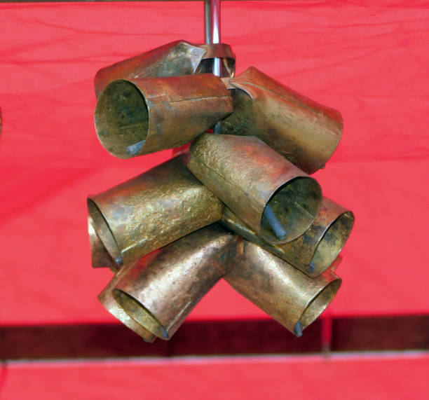 A cluster of metallic cowbells hangs from a central support against a vibrant red background. The bells have a rustic, worn appearance, suggesting they are made of aged brass or bronze. Each bell is cylindrical with a slight flare at the bottom, designed to produce sound when moved. The arrangement is likely decorative or utilitarian for livestock management, emphasizing the traditional use of cowbells in agriculture to locate animals. The background color contrasts sharply with the metallic texture of the bells.