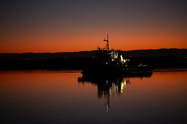 A silhouetted tugboat in calm waters at dusk, with the vibrant orange and deep blue hues of the sunset reflecting on the water