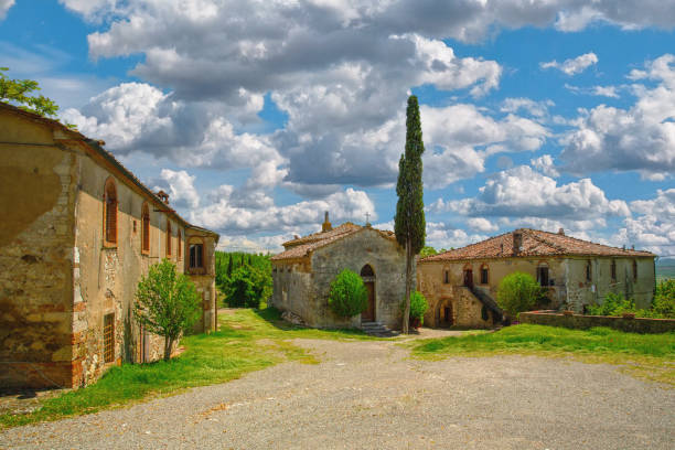 Frosini, a small village in Tuscany, Italy. The scene features a collection of aged stone buildings, including a small chapel with a cross, set against a backdrop of a vibrant blue sky filled with dramatic white clouds. A tall, slender cypress tree stands prominently in the center, adding a vertical element to the composition. The weathered facades of the buildings and the gravel courtyard suggest a long history and a peaceful, rural atmosphere. The lush green grass and scattered trees further enhance the natural beauty of the location.