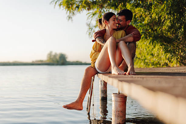 Young couple enjoying a romantic moment on a wooden pier, embracing and dipping their feet in the calm water as the sun sets, creating a peaceful and idyllic scene