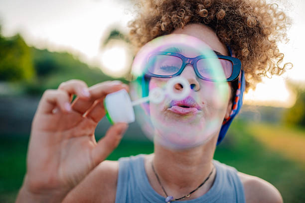 Curly haired young woman wearing stylish glasses is blowing soap bubbles in a park during sunset, embracing the joy of outdoor fun and relishing the carefree spirit of the moment