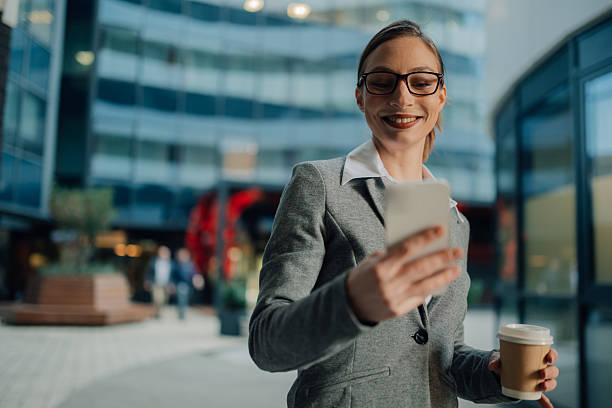 istock Businesswoman using smartphone and holding coffee outside office building 2205454424