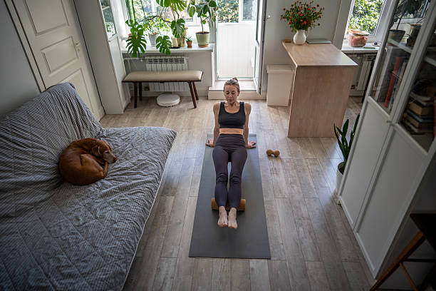 istock Sportive woman breathing deep, do legs exercise with foam roller on mat. Dog napping on couch nearby 2205455196