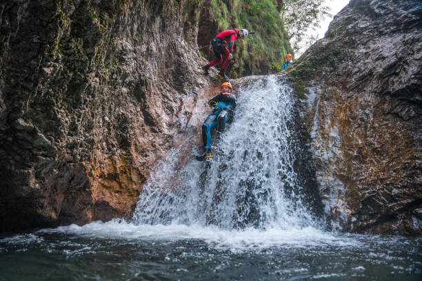 A Caucasian male in a neoprene suit and safety helmet slides down with guidance from a skilled Caucasian male canyoning guide.