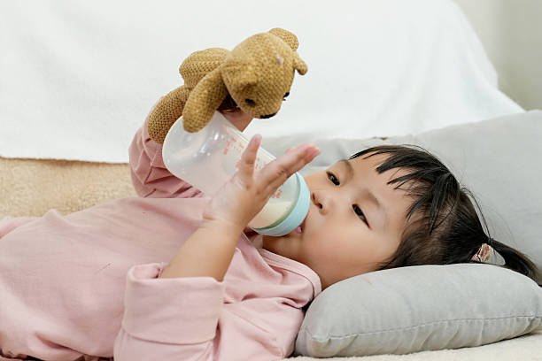 Young daughter lying on sofa drinking milk while holding a stuffed toy. Calm and nurturing home atmosphere showing warmth, love, and the deep connection between a single mother and her child.