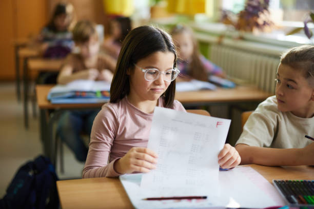 Bad test result at school! Displeased schoolgirl holding a negative test result from her educational exam in the classroom. report card letter f test results education stock pictures, royalty-free photos & images