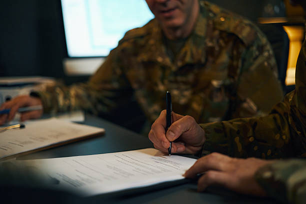 Military personnel signing official documents during work in a dimly lit office environment, focusing on hands and papers, creating a sense of formality and duty