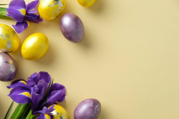 istock Easter flat lay composition. Vibrant golden and purple eggs with purple irises on a light yellow background 2205477422