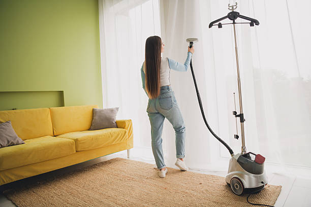 istock Young Woman Using Steam Cleaner on Curtain in Stylish Modern Interior with Comfortable Couch and Daylight 2205484507
