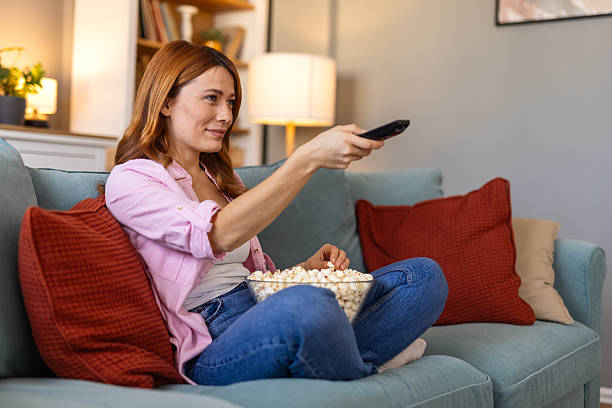 A woman is sitting on a cozy sofa, enjoying a TV show while eating popcorn. The image captures a perfect moment of leisure, ideal for themes related to relaxation, entertainment, and home life.