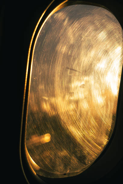 Oval airplane window with a golden glow from sunset light, revealing swirled streaks and scratches across the surface. The light creates a warm, textured appearance, casting subtle reflections inside. The window frame is visible, contrasting with the illuminated view beyond.