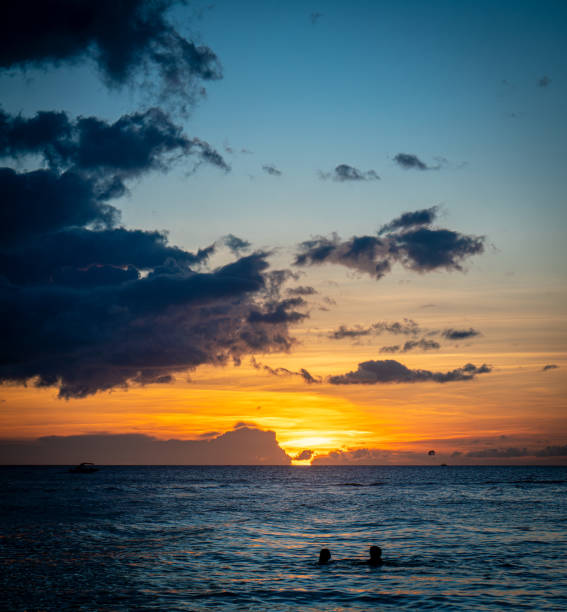 A serene sunset over a calm ocean. Two silhouetted figures float in the foreground, enjoying the tranquil waters. The sky transitions from deep blue at the top to warm hues of orange and yellow near the horizon, with scattered clouds adding depth to the scene. A small distant boat and a parasailing silhouette are visible against the setting sun. This peaceful coastal moment captures the essence of relaxation and natural beauty.