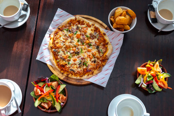 Medium overhead shot of a pizza, potato wedges and salad on a table in a pub in Seghill, Northumberland. The food is surrounded by four coffee cups. A group of people are sharing the food. The pizza is covered with colourful vegetables, making it inviting to eat.\n\nVideos are available similar to this scenario.