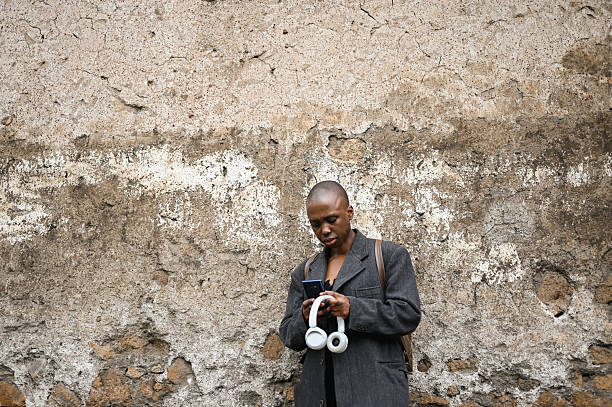 istock Young woman using a smartphone while holding headphones, standing against an old, weathered wall 2205504625