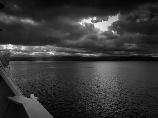 Dramatic black and white seascape with dark clouds and sun rays over calm waters, viewed from a ship deck in New Zealand