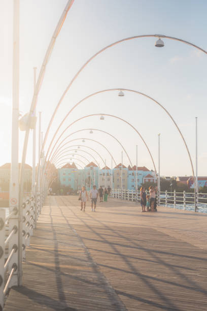 People leisurely strolling across the Queen Emma Bridge, capturing a serene city life scene bathed in warm afternoon light. The vertical shot features a beautiful backlight.