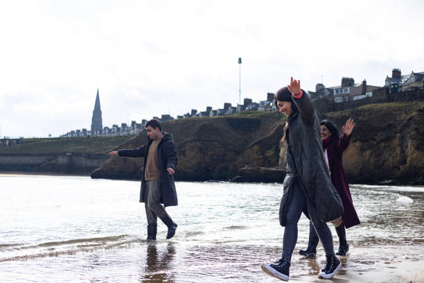 A full shot of a multigenerational family walking along a sandy beach on a cloudy day, dressed in warm coats. The autistic teenage son is enjoying a seaside stroll with his mother and grandmother with scenic cliffs, and houses in the background. Located in Cullerocats, North East of England.\n\nVideos of the same scenario are available