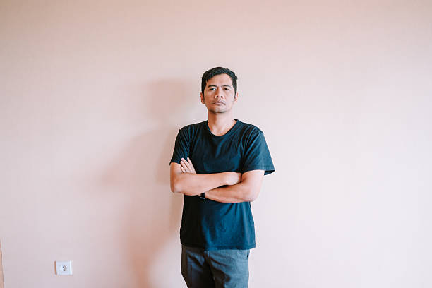 An asian man wearing a dark t-shirt stands confidently with his arms crossed against a plain wall. He has a serious expression, looking directly at the camera. Isolated studio shot, providing ample blank space.