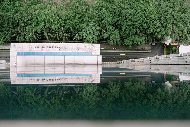 A unique aerial view showcases the harmonious blend of urban architecture and nature. The building's sleek facade is perfectly reflected in a pool of water, mirroring the lush trees and creating a symmetrical and abstract composition. The image captures the essence of contemporary design intertwined with the tranquility of the natural world.