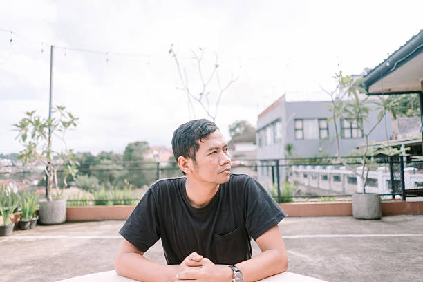 istock Thoughtful asian man looking away while sitting at outdoor rooftop table 2205550279