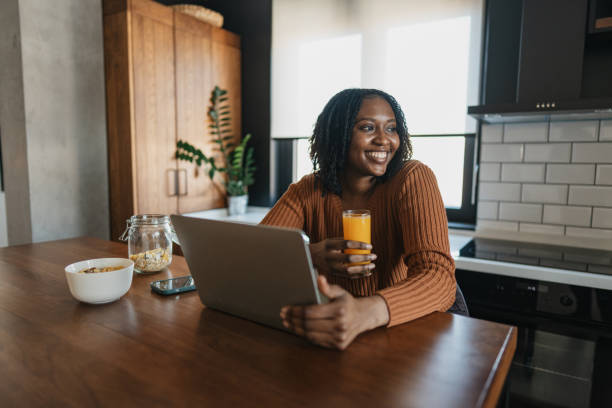 A joyful woman is sitting at a kitchen table with her laptop and a glass of orange juice, embodying a productive and healthy morning routine.