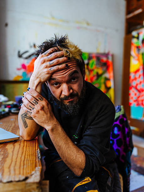 Hispanic man with an alternative look poses thoughtfully in his art studio, surrounded by colorful urban and neo-expressionist artworks.