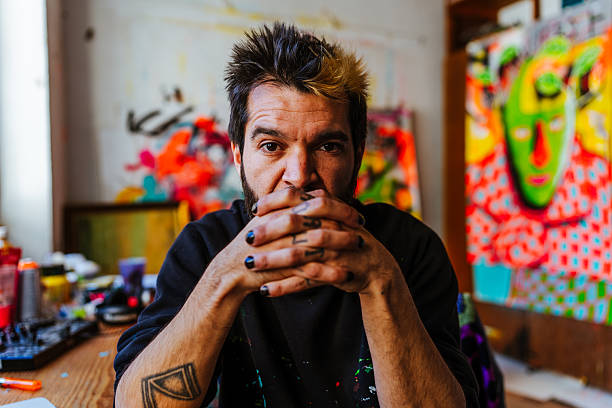 Hispanic Argentine artist with an alternative look poses thoughtfully in his art studio, surrounded by colorful urban and neo-expressionist paintings.