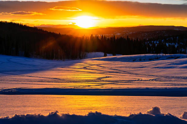 Orange Golden Hour Light Reflecting on Snow with Snowmobile Tracks - Dramatic light with scenic mountain backdrop captured just before sunset with dramatic vivid orange light reflecting on snow surface. Tracks in snow from snowmobiles.