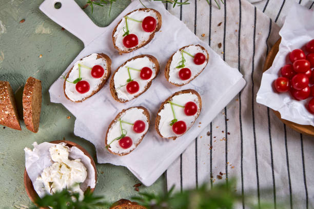 istock Creative tartines with white cheese and tomato cherries rest on a white board on a green table and a striped napkin 2205567482