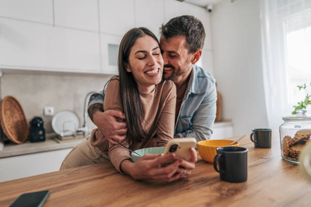 istock Happy couple embracing and using smartphone during breakfast in kitchen 2205587033