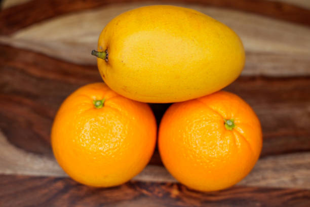A ripe mango placed on top of two fresh oranges, forming a balanced composition on a wooden cutting board. The bright colors contrast beautifully with the warm wood tones.