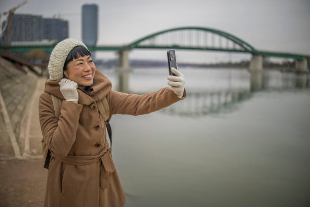 Smiling Japanese woman in warm clothing using mobile phone and having video call  while  standing near river in the city. City life concept  and lifestyle concept.