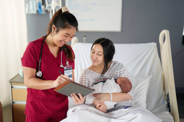 Reviewing Pediatric Testing Results A female pediatric nurse stands in a maternity room at the bedside of a new Mother as she reviews her baby's testing results. She has the results displayed on a tablet as she shares them with the patient who is cradling her swaddled infant in her arms. red medical scrubs stock pictures, royalty-free photos & images