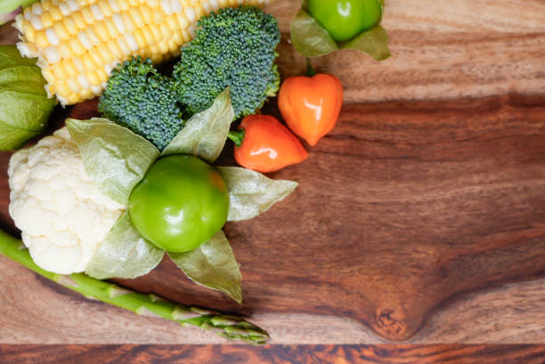 A colorful mix of fresh vegetables, including tomatillo, broccoli, cauliflower, corn, asparagus, and habanero peppers, arranged on a wooden cutting board with a rustic background.