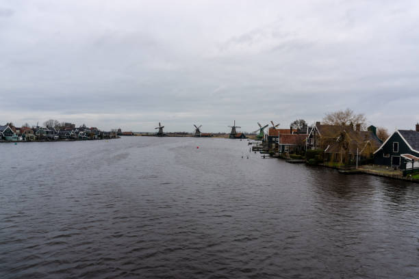 Traditional windmills and charming houses line the riverbanks of zaanse schans in the netherlands, creating a picturesque scene under a cloudy sky