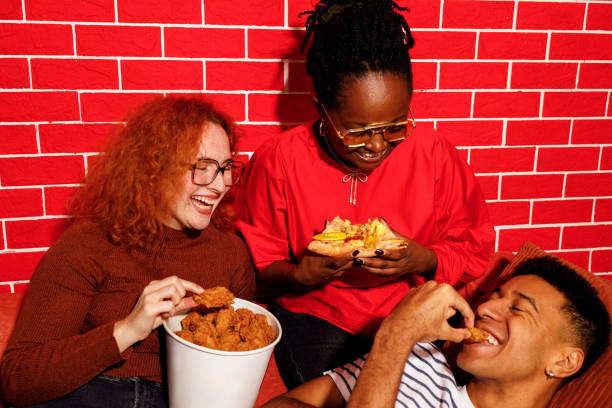 Three young friends, two Black and one red-haired white woman, enjoy a fun moment together while eating different fast food items—pizza, a burger, and chicken wings. They laugh and interact playfully in front of a vibrant red brick wall, capturing the energy and joy of casual dining.